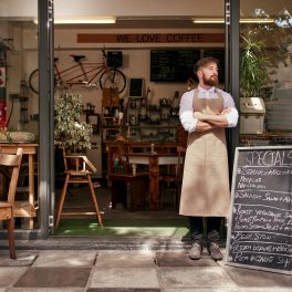 Portrait of a young man standing in front of his coffee shop. Young man with beard wearing an apron  standing with her arms crossed and looking away.